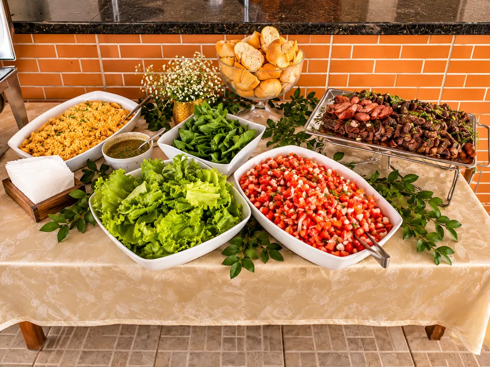 Mesa do buffet com salada de folhas, vinagrete, farofa, pães e espetinhos de carne